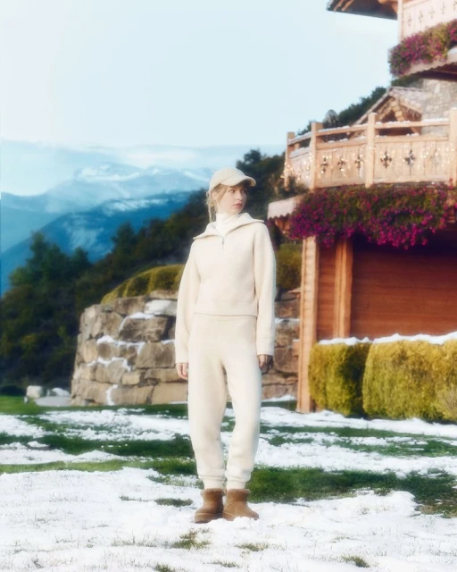 Blonde woman poses outdoors near house wearing beige sweater, sweatpants, cap, and boots in a snowy, mountainous landscape.