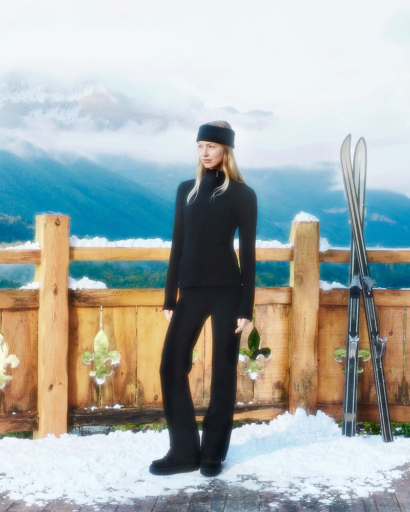 Woman in black ski suit stands near wooden fence with skis in front of snowy mountains on a sunny day, modeling winter apparel.