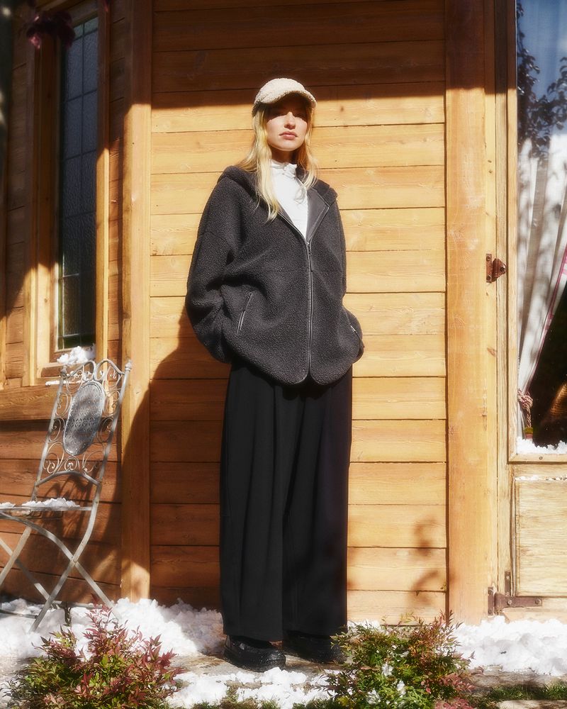 A fair-skinned woman with blonde hair wears a cap, fleece jacket and pants standing outdoors in front of a wooden building.