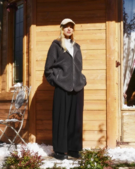 A fair-skinned woman with blonde hair wears a cap, fleece jacket and pants standing outdoors in front of a wooden building.