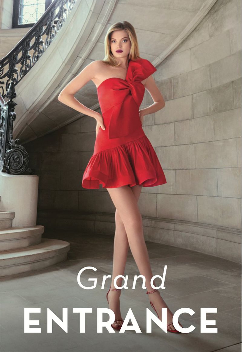 A woman poses in a red dress with a bow on one shoulder, standing on a stone staircase in front of a stone wall and iron railing.