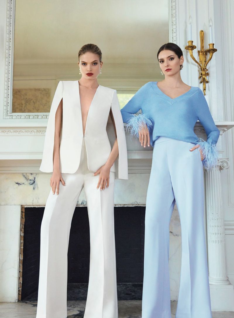 Two women in modern, elegant outfits pose in front of a fireplace with a mirror and wall sconces, conveying a high-fashion aesthetic.