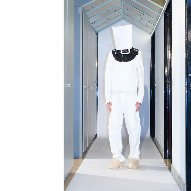 Person in all white clothing with a white paper bag over their head stands in an interior hallway with neutral wall colors.