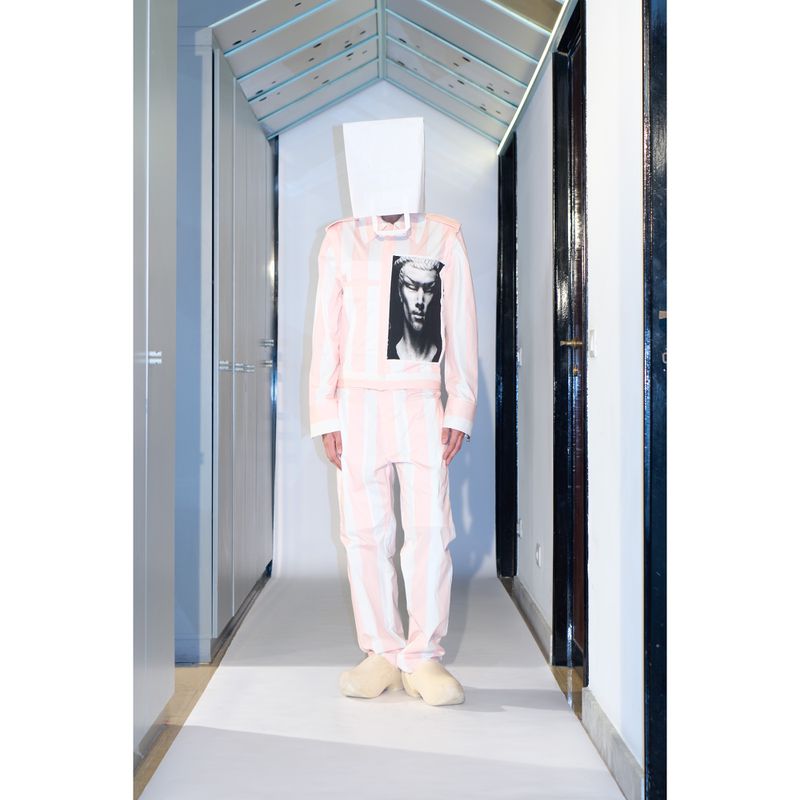 Person in pink and white striped suit with paper bag over head in an indoor hallway with white floors and light blue ceiling.