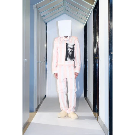 Person in pink and white striped suit with paper bag over head in an indoor hallway with white floors and light blue ceiling.