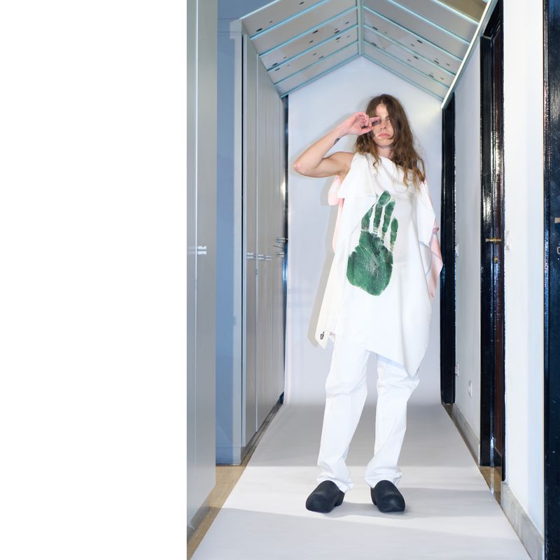 Young adult in white outfit with green handprint stands in a hallway with gray closets and a mirrored ceiling, holding hand up.