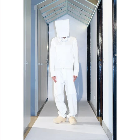 Full-length studio shot of a person in white clothing with a paper bag over their head in a brightly lit hallway.