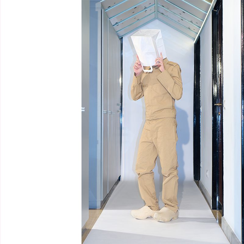 Person in tan outfit poses in hallway with paper bag over head making peace sign with fingers. Hallway is modern, neutral tones.