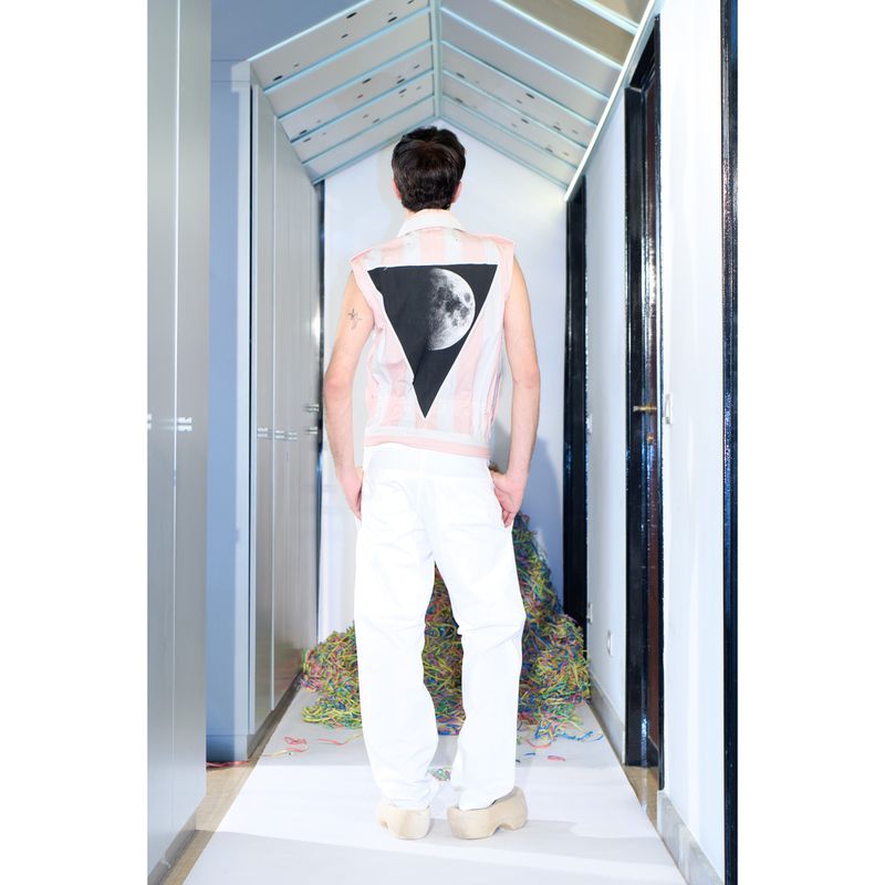Back view of a person wearing a sleeveless top with a moon graphic, white pants, and platform shoes in an indoor setting.