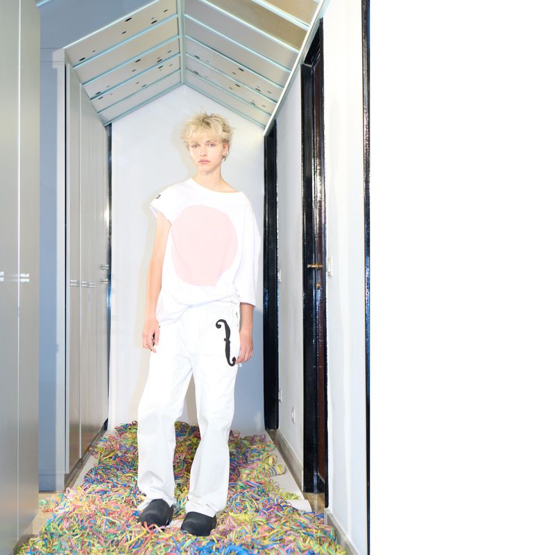 Young adult poses in white streetwear with pastel confetti in indoor setting with reflective ceiling.