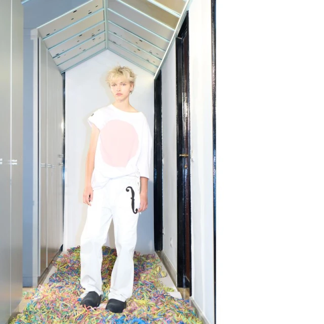 Young adult poses in white streetwear with pastel confetti in indoor setting with reflective ceiling.