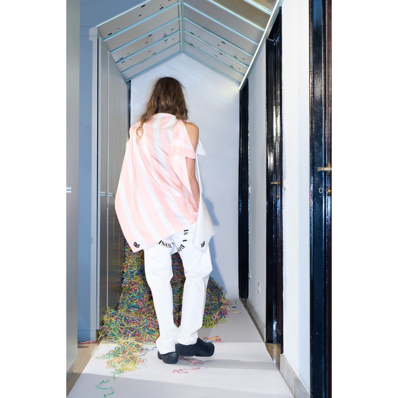 Back view of a young adult wearing a white and pink striped top and white pants stands in a hallway with colorful decorations.
