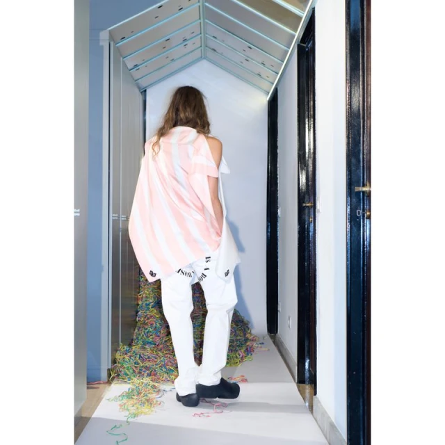 Back view of a young adult wearing a white and pink striped top and white pants stands in a hallway with colorful decorations.