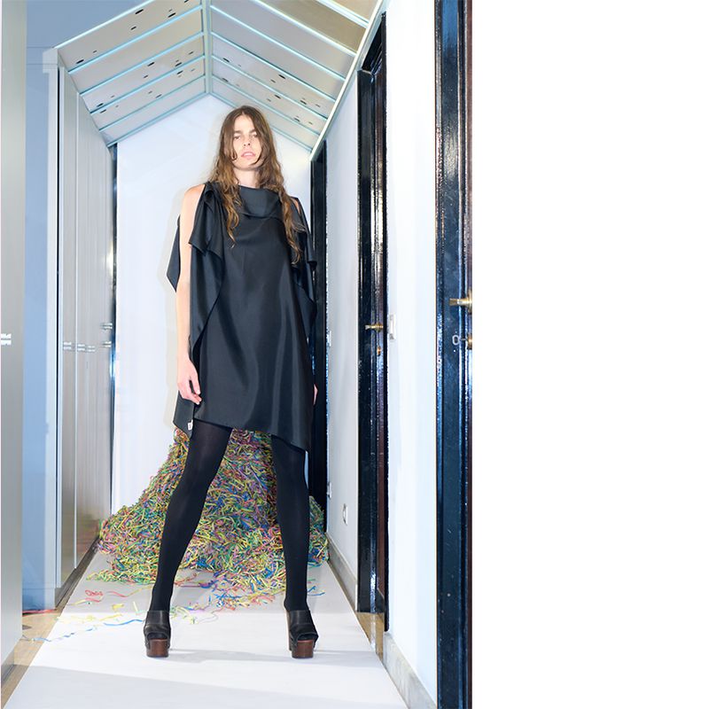 A long-haired woman wearing a black dress, black tights, and platform shoes stands in a narrow hallway with colorful streamers.