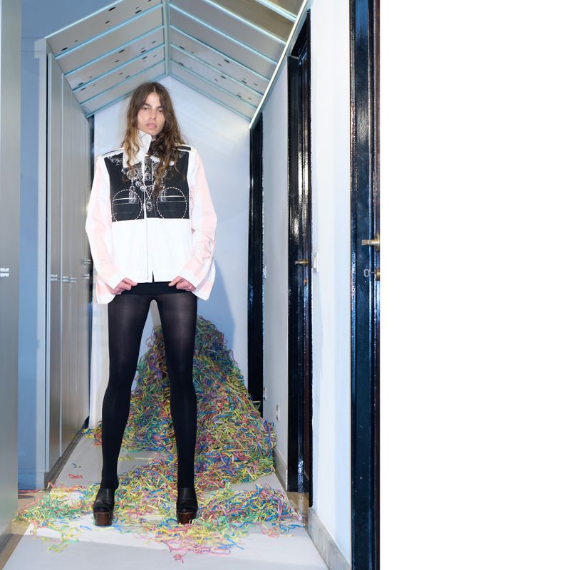 Woman in black tights and platform shoes standing in a hallway with colorful shredded paper on the floor in front of a door.