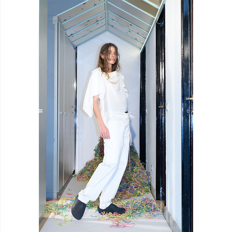 A slender person dressed in a white outfit strikes a fashion pose inside, near a vibrant pile of confetti, with black shoes.