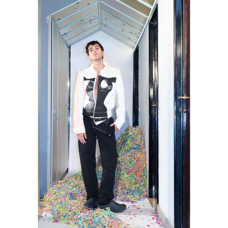 Young adult man in white jacket with black print, black pants, and black shoes, standing indoors amidst a pile of colorful confetti.