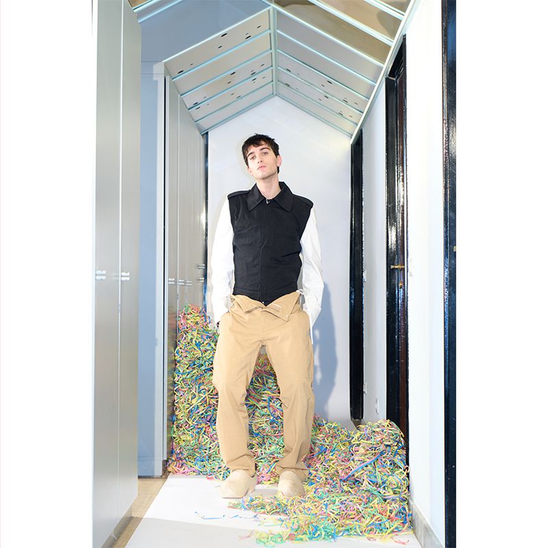 Full-shot of a young man posing in a hallway filled with confetti, wearing a black vest, white long-sleeve shirt and khaki trousers.