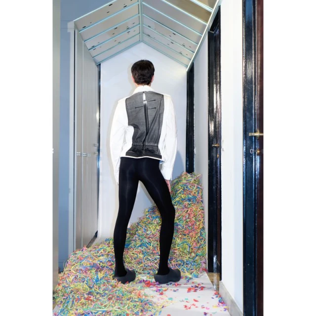 Person stands in an indoor doorway wearing a white button-down shirt, black vest, and black leggings, amidst colorful confetti.