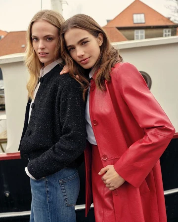 Two women pose together in a medium shot, one in a red coat and the other in a textured jacket, near water and buildings.