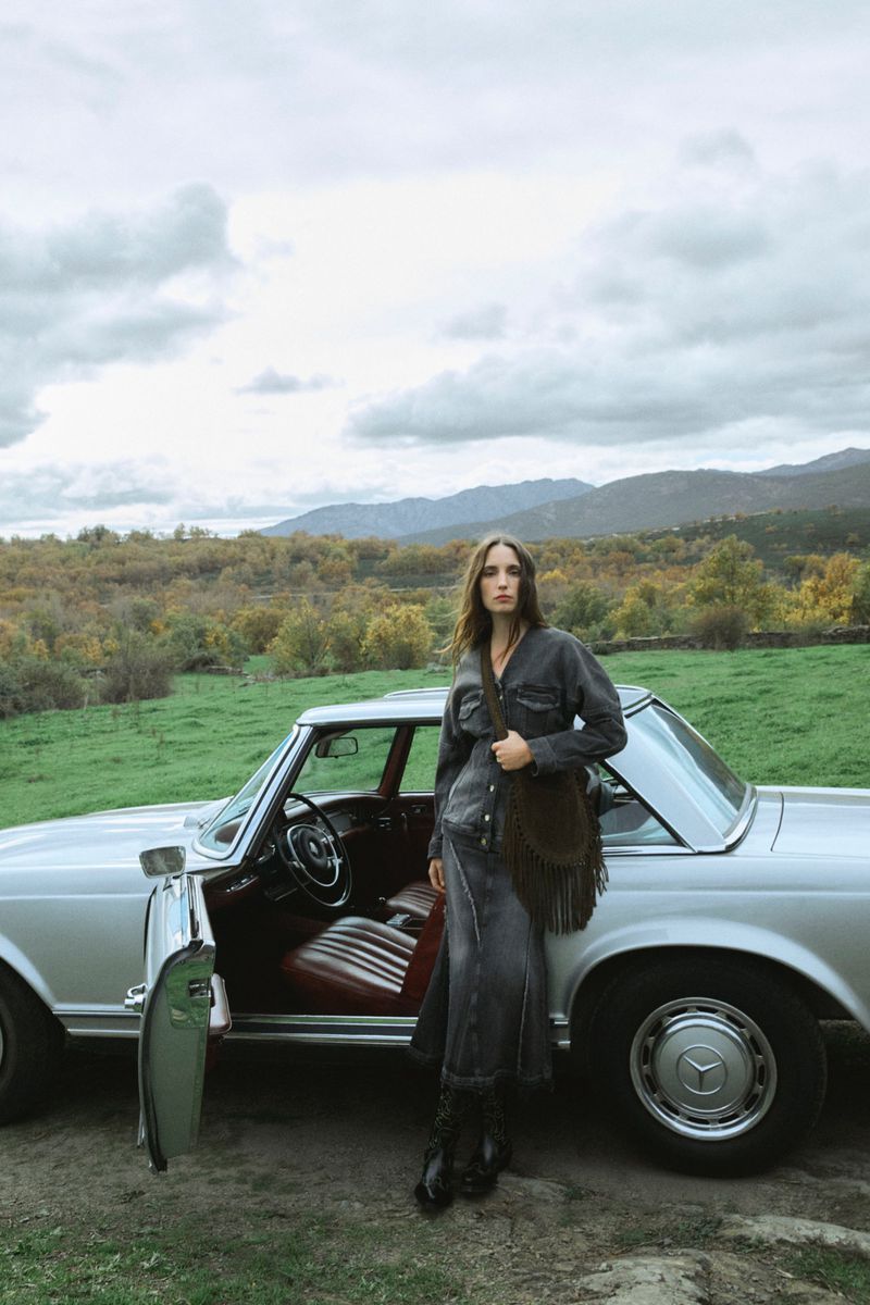 A woman in a denim outfit poses next to a vintage silver car in a rural setting with grassy fields, trees, and cloudy skies.