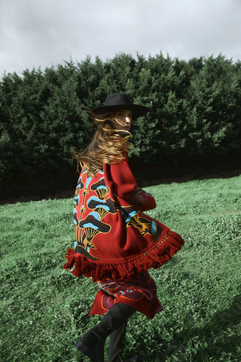 Woman in a red patterned cloak and hat walking in a green grassy field surrounded by trees under a cloudy sky; fashion photography.