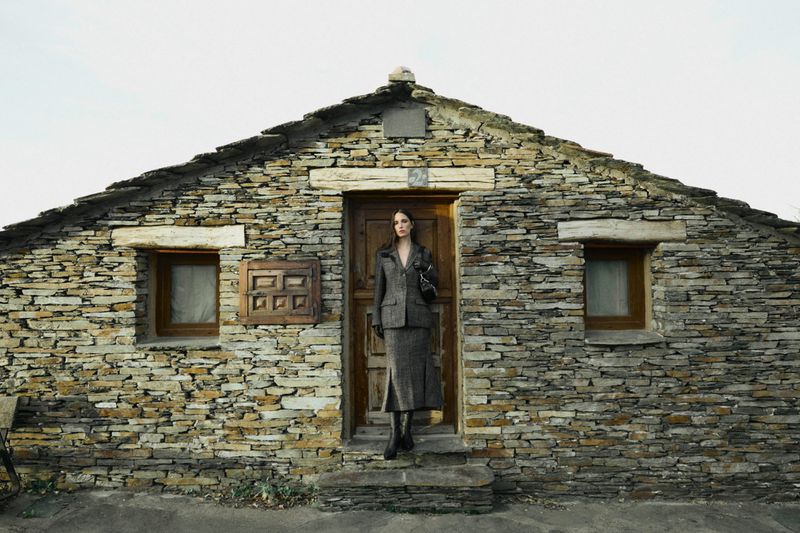 Woman poses in tweed suit in front of stone house with wooden door. Neutral color palette. Fashion editorial photography.