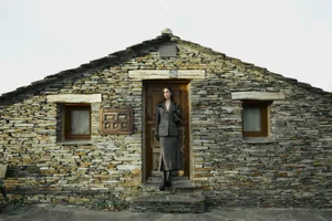 Model standing in the wooden doorway of a rustic stone house wearing a grey patterned skirt suit and dark boots.