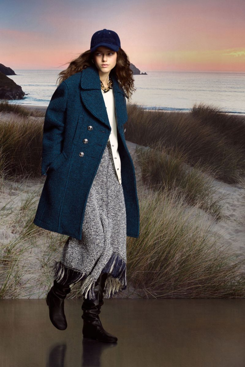 A woman in a blue coat and cap poses on a beach with tall grass and ocean in the background, creating a blend of fashion and nature.