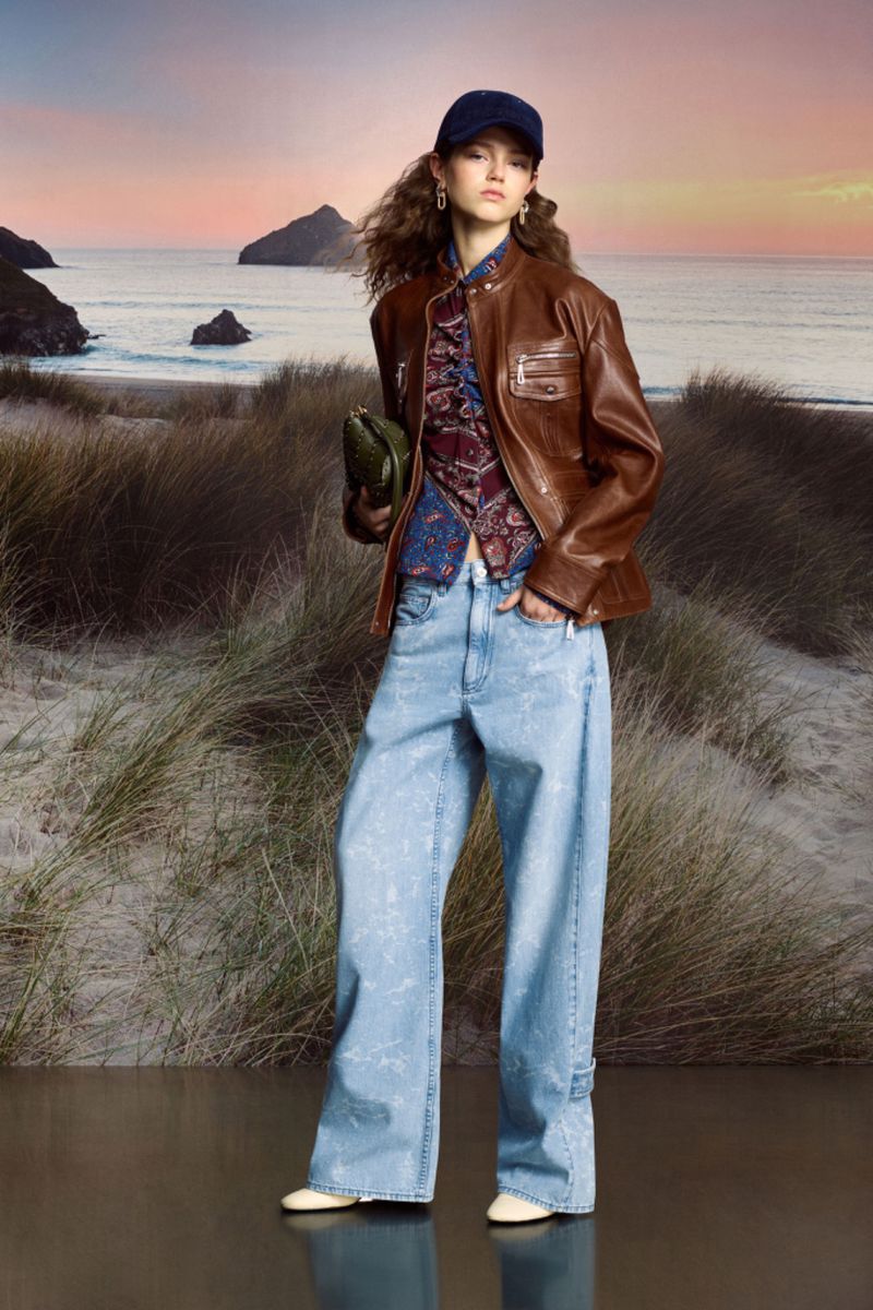 A young woman models wide-leg jeans, a brown leather jacket, and a blue cap on a beach with dunes under a sunset-colored sky.