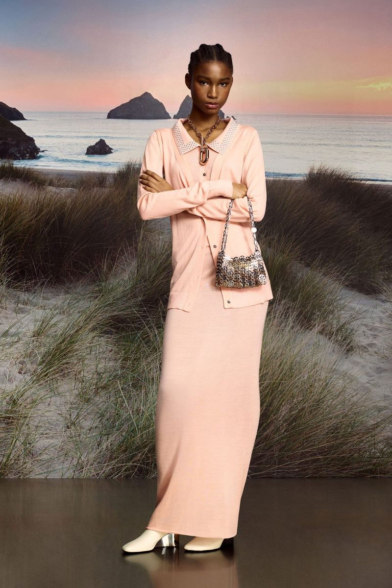 Elegant woman poses in a peach cardigan and maxi skirt on a beach. A small tweed handbag hangs on her shoulder, with sea and sky backdrop.