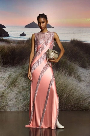 A single woman poses outdoors on a beach at sunset, wearing a formal peach dress with silver trim and holding a clutch bag.