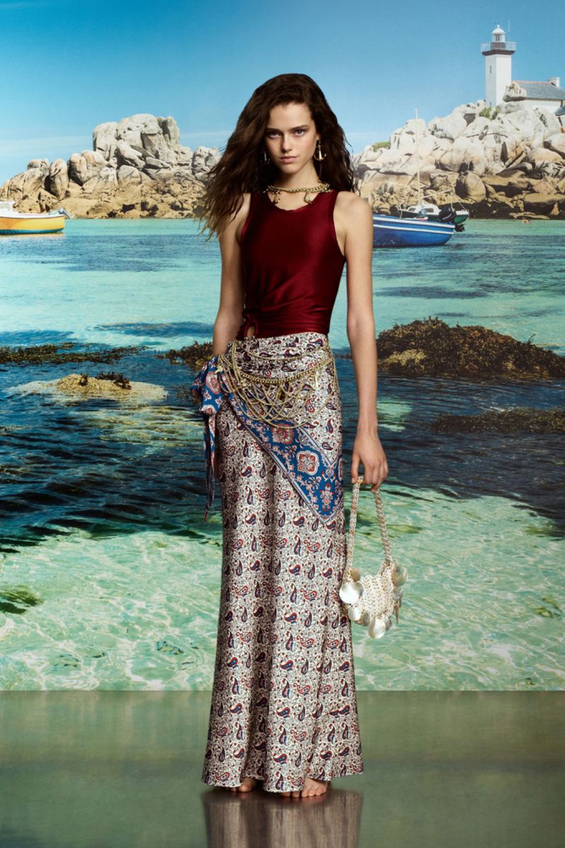 Young woman posing in a maroon top and long printed skirt on a beach with a lighthouse in the background. Fashion shot.