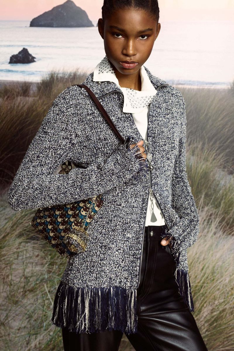 A woman with an intricate handbag wears a textured cardigan, leather pants, and jewelry, posing in front of a beach landscape.