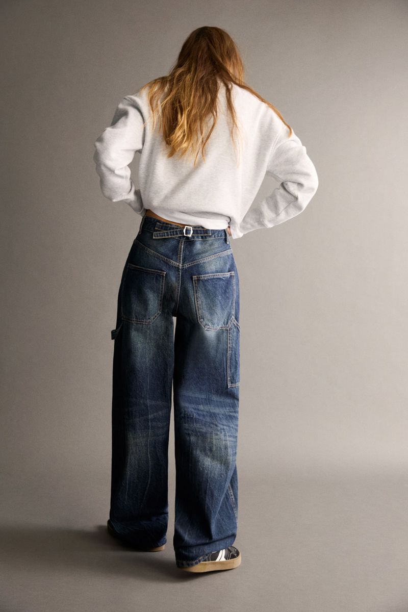 A young woman stands in a gray studio wearing a gray sweatshirt and wide-leg denim carpenter pants with visible pockets and a small belt.