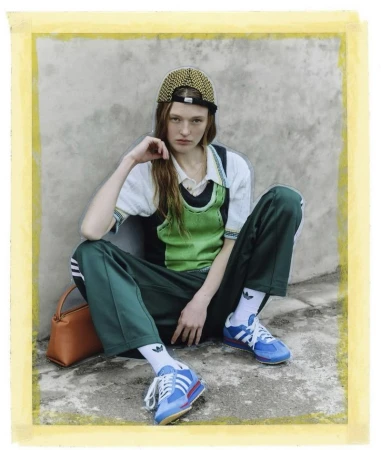 A person sits with green track pants and Adidas sneakers against a grunge wall. The model has long reddish-brown hair and a cap.