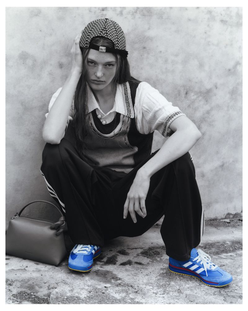 Monochrome fashion shot of a female model crouching against a wall, wearing a patterned cap, top, vest, track pants, and Adidas sneakers.