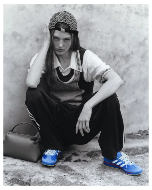 Monochrome fashion shot of a female model crouching against a wall, wearing a patterned cap, top, vest, track pants, and Adidas sneakers.