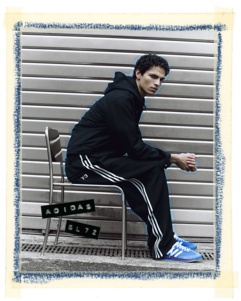 A male model sits on a chair in front of a corrugated metal wall, wearing a black Adidas tracksuit and blue Adidas sneakers, with Adidas logo text.