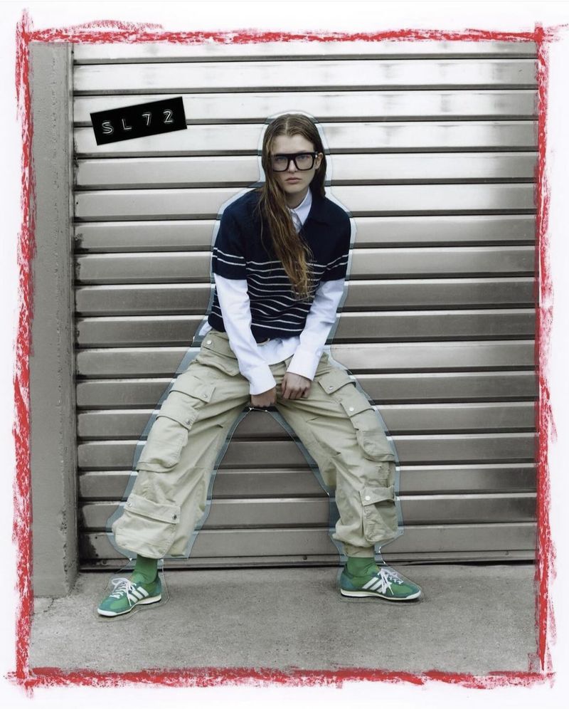 A young adult woman poses against a metal garage door, wearing streetwear fashion including green sneakers and large black glasses.