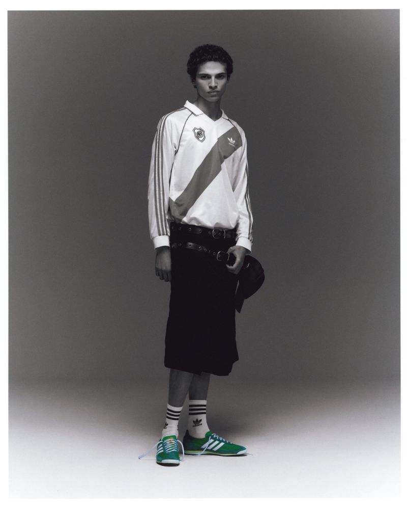 A person stands in a photo studio wearing a white soccer jersey, black skirt, white socks, and green sneakers against a gray background.