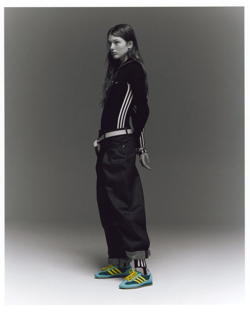 A slender young woman in a black Adidas track jacket and baggy black jeans poses indoors against a neutral backdrop, wearing blue and yellow sneakers.