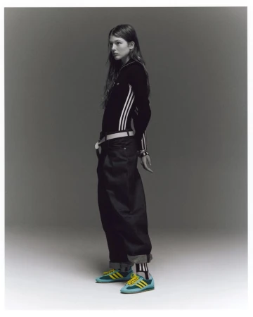 A slender young woman in a black Adidas track jacket and baggy black jeans poses indoors against a neutral backdrop, wearing blue and yellow sneakers.