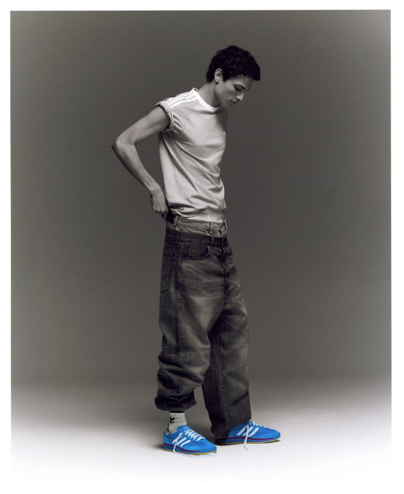 Young man in a white t-shirt and baggy jeans stands in a photo studio with blue Adidas sneakers, looking down. Fashion style photography.