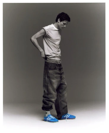 Young man in a white t-shirt and baggy jeans stands in a photo studio with blue Adidas sneakers, looking down. Fashion style photography.