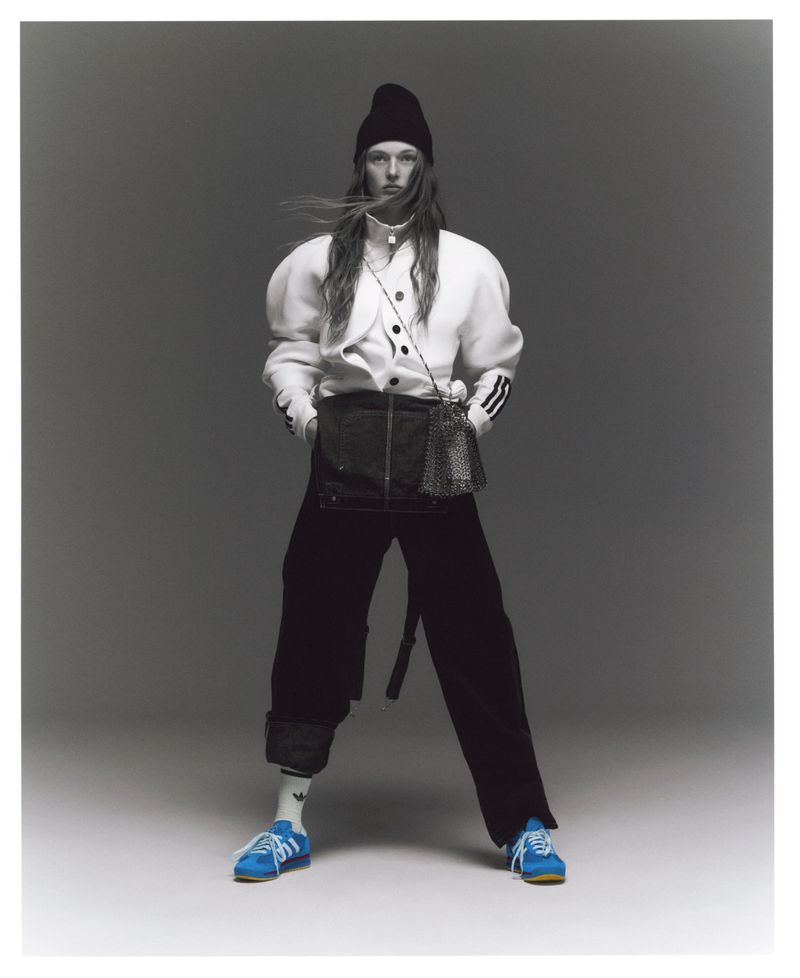 A full-length studio shot of a non-binary model wearing a black beanie, puffy-sleeve white shirt, black skirt, black pants, and blue sneakers.