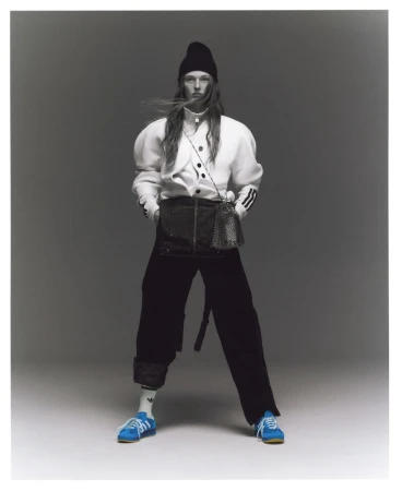 A full-length studio shot of a non-binary model wearing a black beanie, puffy-sleeve white shirt, black skirt, black pants, and blue sneakers.
