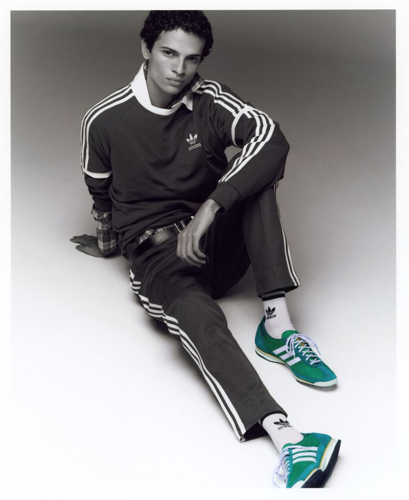An adult male model poses in a studio wearing a burgundy Adidas tracksuit, white socks with the Adidas logo, and green Adidas sneakers.