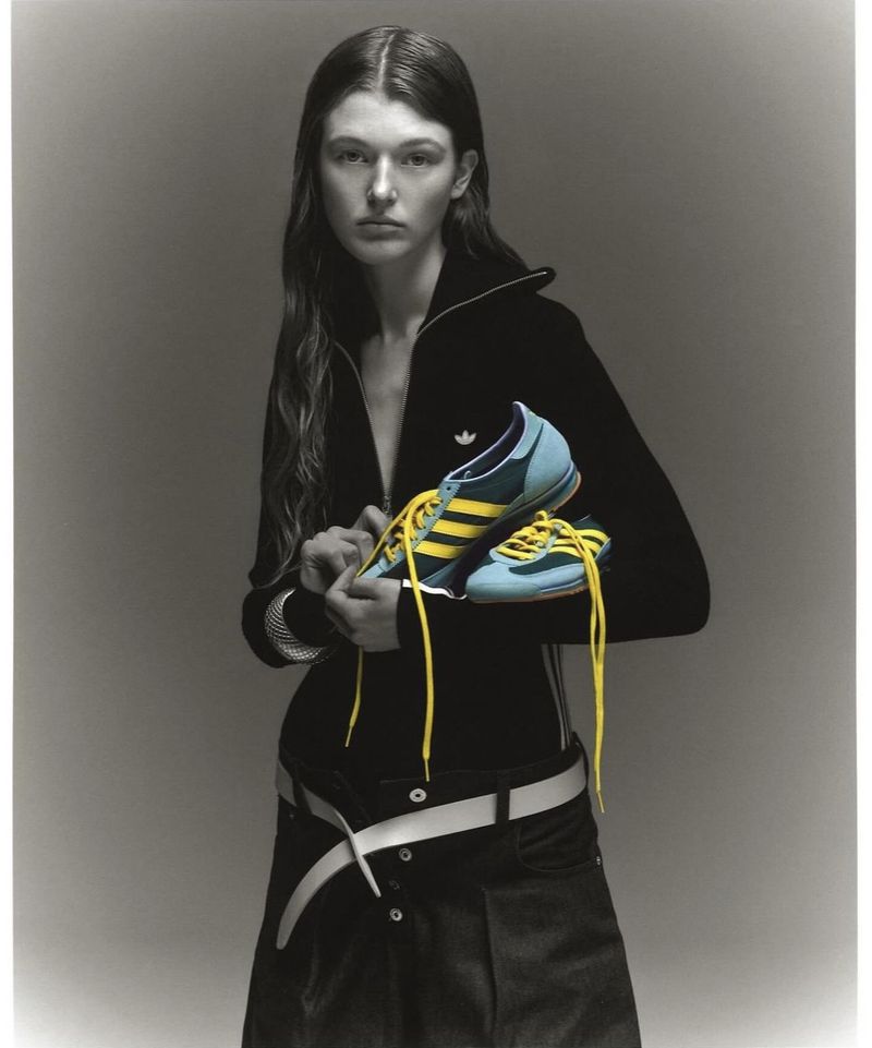 Young woman poses in a black athletic jacket holding blue and yellow Adidas shoes in a studio with neutral grey backdrop.