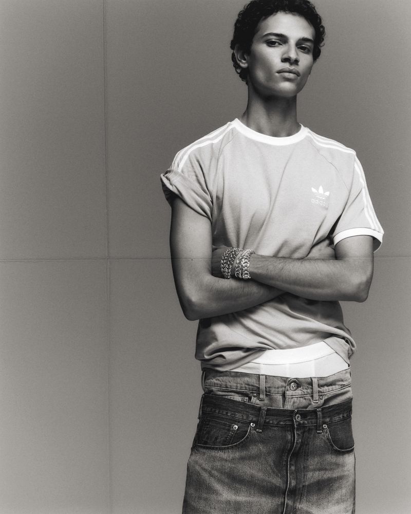 Black-and-white portrait of an adult male with curly hair, wearing an Adidas T-shirt and jeans, posing with arms crossed indoors.